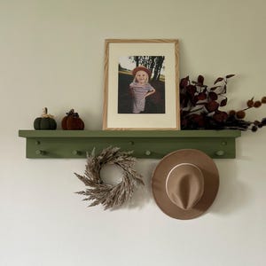 May include: A green shelf with a framed photo of a child wearing a red hat, two small pumpkins, a dried grass wreath, and a brown fedora hat hanging on hooks.