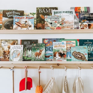 Pu&ograve; includere: Uno scaffale in legno con una variet&agrave; di libri per bambini, tra cui titoli come "The Tale of Peter Rabbit", "Old Rock", "Seasons" e "Love You Baby". Lo scaffale &egrave; montato su una parete con ganci sotto per appendere oggetti.