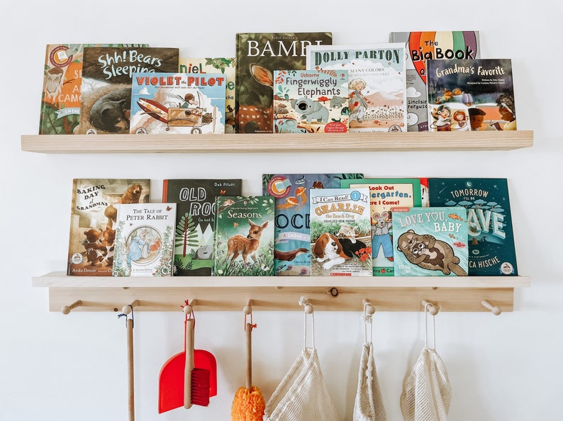 Pu&ograve; includere: Uno scaffale in legno con una variet&agrave; di libri per bambini, tra cui titoli come "The Tale of Peter Rabbit", "Old Rock", "Seasons" e "Love You Baby". Lo scaffale &egrave; montato su una parete con ganci sotto per appendere oggetti.