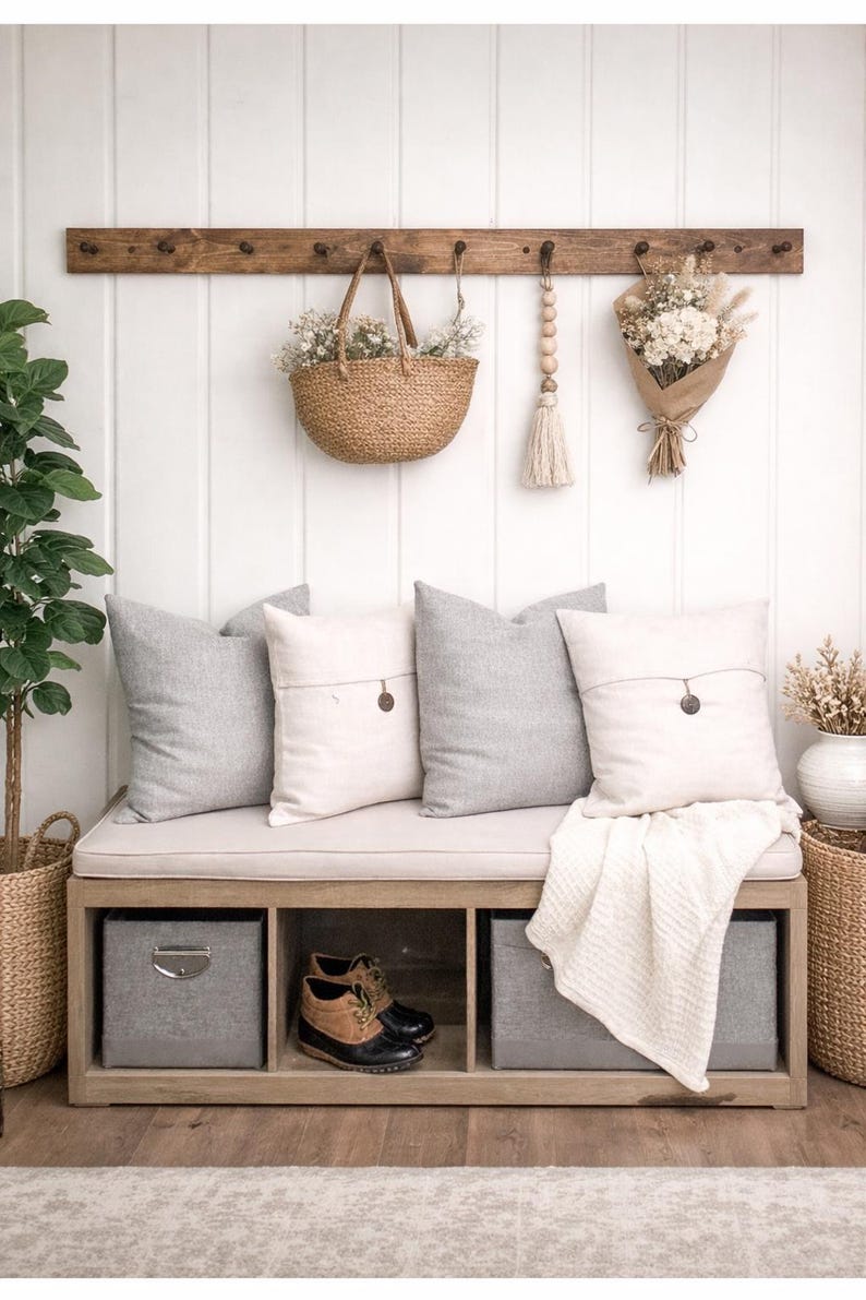 May include: A wooden bench with gray storage bins and a cream-colored cushion is adorned with gray and white pillows. A woven basket and a bouquet of dried flowers hang above. A pair of brown and black boots sits in the bench's open compartment.