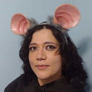 以下が含まれることがあります： 大きなピンク色のネズミの耳が付いたグレーのヘアバンド。耳はリアルで質感のある外観をしています。ヘアバンドはグレーのふわふわの毛皮で飾られています。画像内の人物は黒いパーカーを着ています。