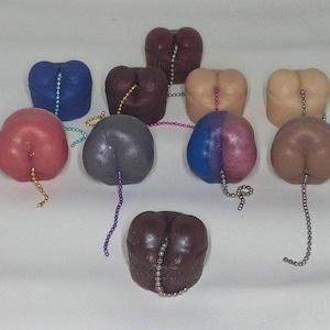 Puede incluir: Una colección de artículos novedosos con forma de trasero en varios colores, incluyendo rojo, azul y marrón. Cada artículo tiene una pequeña cadena adjunta. Los artículos están dispuestos sobre una superficie blanca.