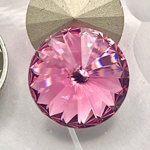 Peut inclure: Un bouton rond en cristal rose à facettes avec un motif en étoile, placé à côté d'une pièce de monnaie argentée. Le bouton présente un dos métallique texturé de forme conique, affichant une teinte rose translucide vibrante et des reflets lumineux géométriques nets sur sa surface.