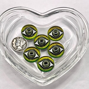 May include: Seven round glass eye cabochons with a black iris, white eyelids, and yellow and green accents. A silver coin is next to the cabochons in a clear heart-shaped dish. The cabochons are approximately 0.75 inches in diameter.