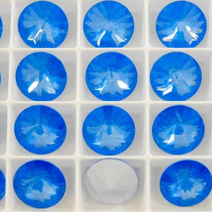 May include: A tray of round, faceted, blue crystal rhinestones. Each gem is set in a white square compartment. One compartment contains a white crystal. The gems have a starburst pattern.