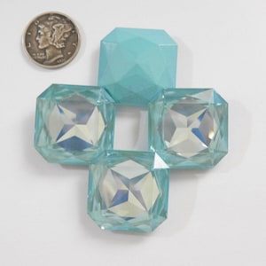 以下が含まれることがあります： 銀貨と、4つの水色のファセット加工されたクリスタルのようなオブジェクトが十字型に配置されています。オブジェクトは幾何学的なデザインで、光を反射します。コインには横顔が描かれています。