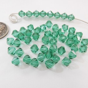 May include: A scattering of small, faceted, green glass beads. The beads are in the shape of a pyramid with a pointed top. The beads are scattered on a white surface.