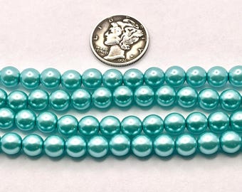 May include: Three strands of light turquoise faux pearls are displayed next to a silver coin. The pearls are round and have a glossy finish. The coin is a silver dime from 1936.