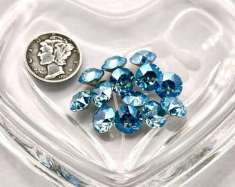May include: A collection of light blue faceted gemstones in a clear, heart-shaped glass dish, next to a silver coin. The gemstones vary in size and shape, reflecting light. The coin is a 1936 Mercury dime.