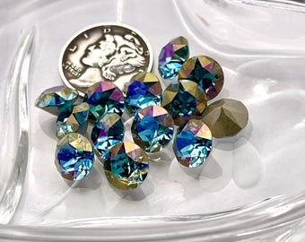 Può includere: Una collezione di gemme iridescenti sfaccettate nei toni del blu, verde e oro, esposte in una ciotola di vetro trasparente. È presente anche una moneta d'argento. Le gemme variano per forma e dimensione, riflettendo la luce.