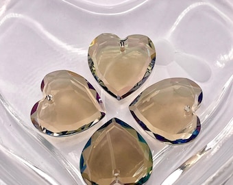 May include: Four heart-shaped crystal pendants with iridescent edges rest in a clear, heart-shaped glass dish. The pendants have a hole at the top for stringing. The crystals reflect light, displaying a range of colors.