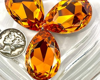 Peut inclure: Trois pierres précieuses orange en forme de goutte d'eau, taillées à facettes avec une coupe brillante, présentées sur un plat en verre transparent en forme de cœur. Une pièce de monnaie en argent est également présente. Les pierres ont une teinte ambrée chaude.