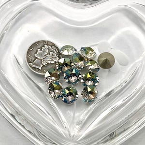 May include: A collection of iridescent crystal rhinestones and a silver coin are displayed in a heart-shaped glass dish. The rhinestones have a shimmering, multi-colored effect. The coin is a Mercury dime from 1936.