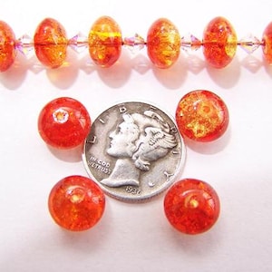 Puede incluir: Una colección de cuentas de vidrio naranja y amarillo, algunas redondas y otras ovaladas, con una moneda de plata. Las cuentas están ensartadas en un alambre y dispersas alrededor de la moneda. Las cuentas tienen un aspecto moteado y texturizado.