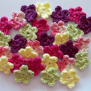 Könnte beinhalten: Eine Ansammlung gehäkelter Blumenapplikationen in verschiedenen Farben, darunter Rosa, Lila, Gelb und Grün. Die handgefertigten Blumen sind auf einer weißen Oberfläche angeordnet und zeigen ihr detailliertes Blütenblattdesign. Diese floralen Verzierungen eignen sich für Bastelarbeiten.