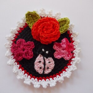 Pode incluir: Uma aplicação de flores de crochê com uma rosa vermelha, flores cor-de-rosa, uma joaninha e uma borda branca de crochê.