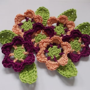 Peut inclure: Un groupe de fleurs crochetées dans des tons de rose, vert et violet. Les fleurs sont disposées en cercle et ont un aspect délicat et ajouré.