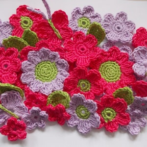 Puede incluir: Una colección de flores de ganchillo en tonos de rosa, morado y verde. Las flores son de diferentes tamaños y formas, y están dispuestas en un patrón aleatorio.
