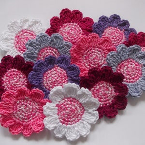 Op de afbeelding: Een verzameling van tien gehaakte bloemen in verschillende tinten roze, wit, paars en grijs. De bloemen zijn allemaal ongeveer even groot en hebben een vergelijkbaar ontwerp.