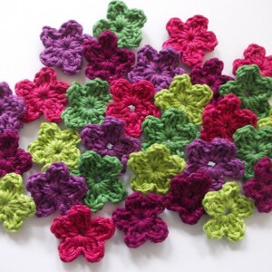 Op de afbeelding: Een verzameling gehaakte bloemenversieringen in tinten groen, paars en roze. De bloemen zijn allemaal vijfpuntige sterren met een centraal gat.