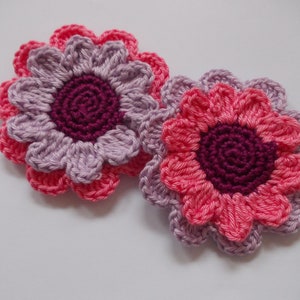 Op de afbeelding: Twee gehaakte bloemen met roze en paarse bloemblaadjes en een bordeauxrode kern.