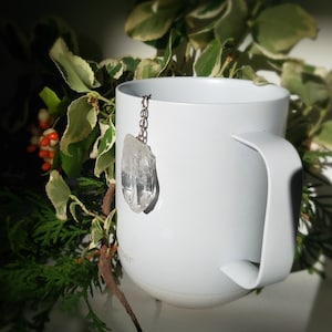 May include: A white ceramic mug with a crystal charm hanging from the handle. The mug is sitting on a table with green plants in the background.