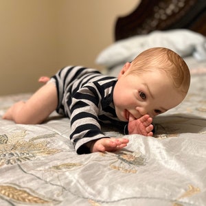 Puede incluir: Un mu&ntilde;eco de beb&eacute; con un mono a rayas est&aacute; acostado boca abajo en una cama con un edred&oacute;n estampado floral. El mu&ntilde;eco est&aacute; mirando hacia abajo y tiene su mano en su boca.