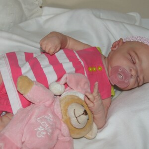 May include: A pink and white striped dress with yellow buttons, a pink pacifier, and a pink teddy bear with the text "We Love Me" on it. The baby doll is wearing a pink headband with a pink flower.