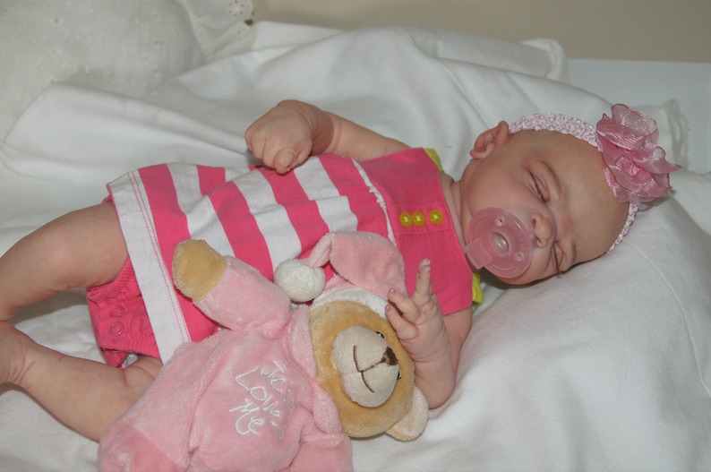 May include: A pink and white striped dress with yellow buttons, a pink pacifier, and a pink teddy bear with the text "We Love Me" on it. The baby doll is wearing a pink headband with a pink flower.