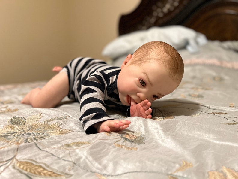 Puede incluir: Un mu&ntilde;eco de beb&eacute; con un mono a rayas est&aacute; acostado boca abajo en una cama con un edred&oacute;n estampado floral. El mu&ntilde;eco est&aacute; mirando hacia abajo y tiene su mano en su boca.