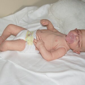 May include: A realistic baby doll wearing a diaper and a pink pacifier, lying on a white sheet.