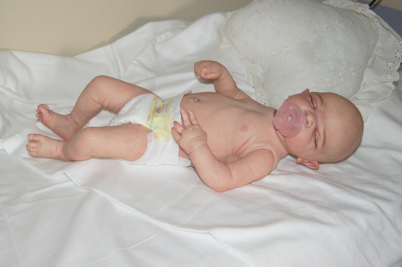 May include: A realistic baby doll wearing a diaper and a pink pacifier, lying on a white sheet.