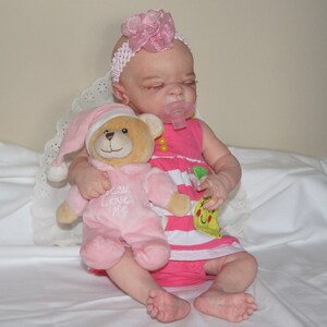 May include: A pink and white baby doll with a pink flower headband is holding a pink teddy bear with the text "Can Love Me" on its chest. The doll is wearing a pink and white striped dress with a yellow and green fruit design.