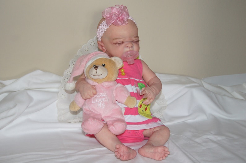 May include: A pink and white baby doll with a pink flower headband is holding a pink teddy bear with the text "Can Love Me" on its chest. The doll is wearing a pink and white striped dress with a yellow and green fruit design.