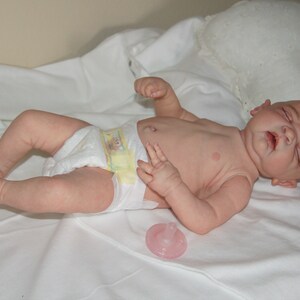 May include: A realistic baby doll with closed eyes, wearing a diaper and lying on a white sheet. A pink pacifier is on the sheet next to the doll.