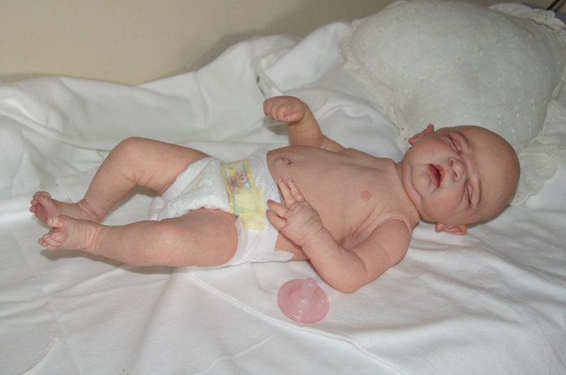 May include: A realistic baby doll with closed eyes, wearing a diaper and lying on a white sheet. A pink pacifier is on the sheet next to the doll.