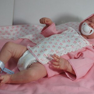 May include: A pink and white baby doll wearing a pink and white floral print onesie with a white pacifier in its mouth. The doll is lying on a pink blanket.