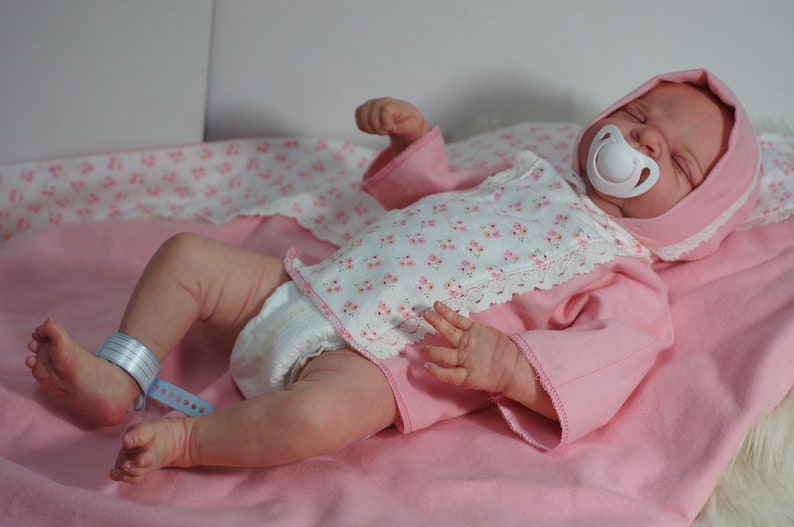 May include: A pink and white baby doll wearing a pink and white floral print onesie with a white pacifier in its mouth. The doll is lying on a pink blanket.