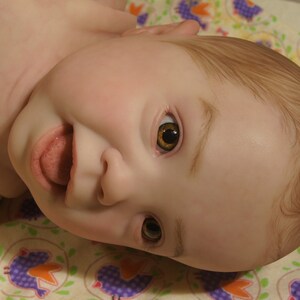 Puede incluir: Primer plano de una mu&ntilde;eca beb&eacute; con cabello casta&ntilde;o y ojos abiertos. La mu&ntilde;eca est&aacute; acostada boca arriba con la boca abierta y la lengua fuera. La mu&ntilde;eca lleva un atuendo de color marr&oacute;n claro.