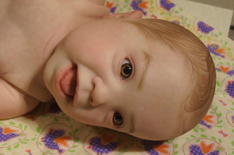 Puede incluir: Primer plano de una mu&ntilde;eca beb&eacute; con cabello casta&ntilde;o y ojos abiertos. La mu&ntilde;eca est&aacute; acostada boca arriba con la boca abierta y la lengua fuera. La mu&ntilde;eca lleva un atuendo de color marr&oacute;n claro.