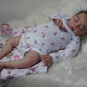 May include: A sleeping baby doll with closed eyes, wearing a white long-sleeved onesie with a pink and blue owl pattern. The doll is lying on a white fluffy blanket with a pink and white patterned fabric underneath.