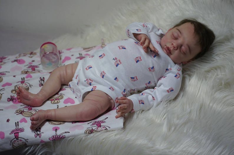 May include: A sleeping baby doll with closed eyes, wearing a white long-sleeved onesie with a pink and blue owl pattern. The doll is lying on a white fluffy blanket with a pink and white patterned fabric underneath.