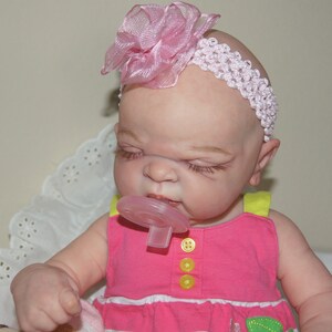 May include: A pink and white headband with a pink flower on top is worn by a baby doll with closed eyes and a pacifier in its mouth. The doll is wearing a pink and yellow dress.
