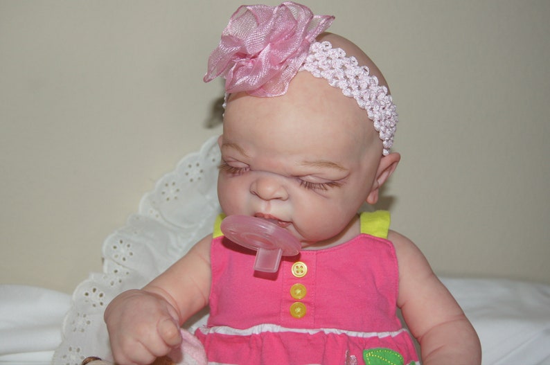 May include: A pink and white headband with a pink flower on top is worn by a baby doll with closed eyes and a pacifier in its mouth. The doll is wearing a pink and yellow dress.