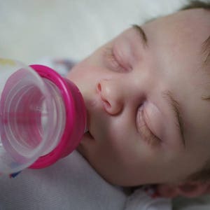 May include: A baby is sleeping while being fed from a clear plastic bottle with a pink and white nipple.