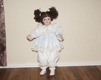 Puede incluir: Una muñeca de porcelana con cabello castaño rizado peinado en dos moños altos, vestida con un vestido azul claro y blanco con ribete de encaje. La muñeca está de pie sobre un piso de madera.