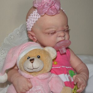 May include: A baby doll wearing a pink dress and a white headband with a pink flower. The doll is holding a pink teddy bear with a white hat. The doll is also wearing a pink pacifier.