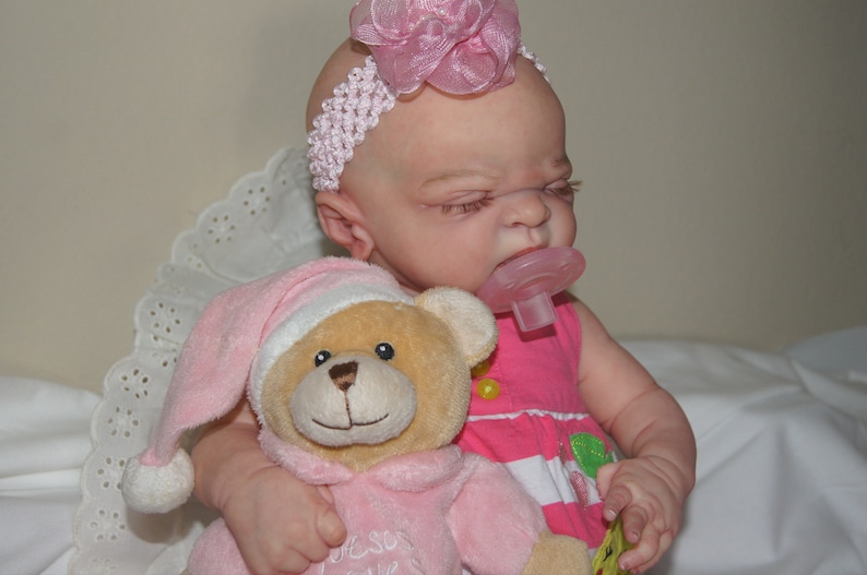 May include: A baby doll wearing a pink dress and a white headband with a pink flower. The doll is holding a pink teddy bear with a white hat. The doll is also wearing a pink pacifier.