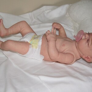 May include: A realistic baby doll with closed eyes, wearing a diaper and a pink pacifier. The doll is lying on a white sheet.