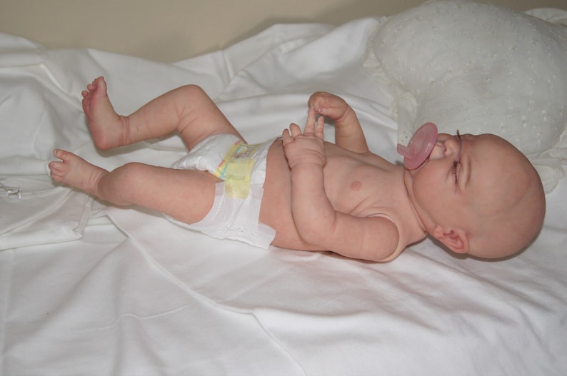 May include: A realistic baby doll with closed eyes, wearing a diaper and a pink pacifier. The doll is lying on a white sheet.